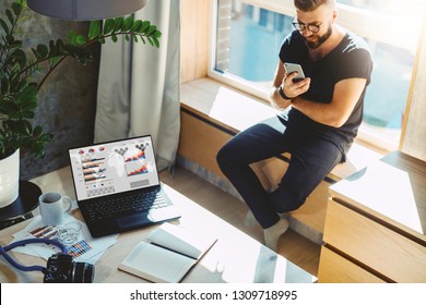 Handsome Hipster Man Sits Home On Windowsill, Reads Text Messages, Sends Email, Uses His Phone And Fast Internet Connection. On Table Laptop With Graphs, Charts, Diagrams On Monitor, Camera, Notebook.