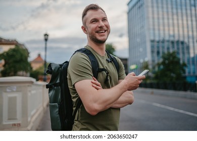 handsome hipster man walking in street with urban backpack traveler in khaki colors, using smartphone, smiling happy