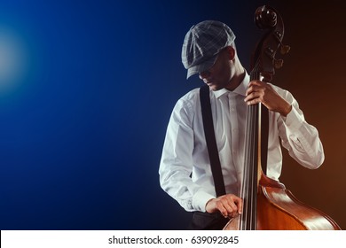 Handsome hipster musician playing bass, close-up. Wearing plaid flat cap, white shirt and suspenders, concert lights. Jazz festival.