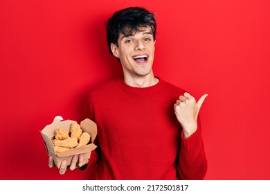 Handsome Hipster Young Man Eating Chicken Wings Pointing Thumb Up To The Side Smiling Happy With Open Mouth 