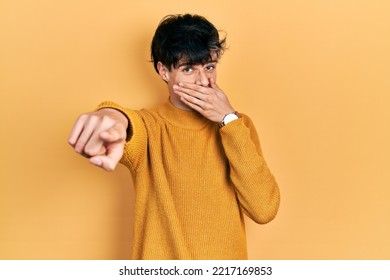 Handsome Hipster Young Man Wearing Casual Yellow Sweater Laughing At You, Pointing Finger To The Camera With Hand Over Mouth, Shame Expression 