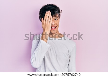 Handsome hipster young man wearing casual white shirt covering one eye with hand, confident smile on face and surprise emotion. 