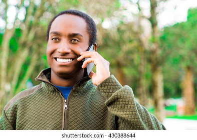 Handsome Hispanic Black Man Wearing Green Sweater In Outdoors Park Area Holding Up Phone To Ear And Talking Happily While Laughing