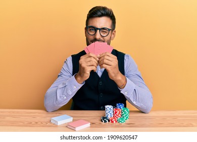 Handsome hispanic croupier man sitting on the table with poker chips and cards winking looking at the camera with sexy expression, cheerful and happy face. 