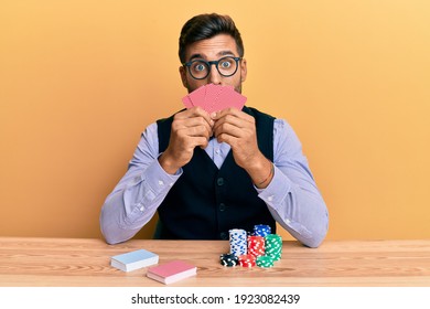 Handsome hispanic croupier man sitting on the table with poker chips and cards in shock face, looking skeptical and sarcastic, surprised with open mouth 