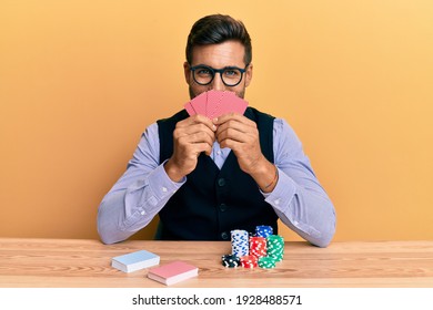 Handsome hispanic croupier man sitting on the table with poker chips and cards smiling with a happy and cool smile on face. showing teeth. 