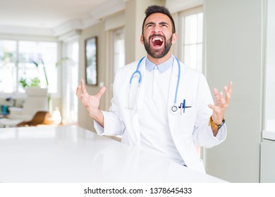 Handsome hispanic doctor man wearing stethoscope at the clinic crazy and mad shouting and yelling with aggressive expression and arms raised. Frustration concept. - Shutterstock ID 1378645433
