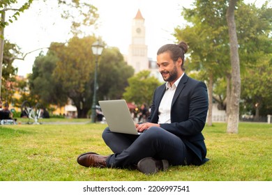 Handsome Hispanic Male Businessman Sitting Lotus Position In Public Park Use Laptop. Remote Work Concept. Stylish European Man In Suit Relax In Park Summer Day Working On Computer. Technology.