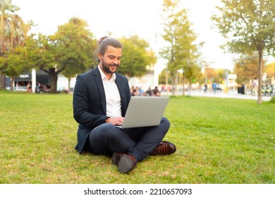 Handsome Hispanic Male Businessman Sitting Lotus Position In Public Park Use Laptop. Remote Work Concept. Stylish European Man In Suit Relax In Park Summer Day Working On Computer. Technology.