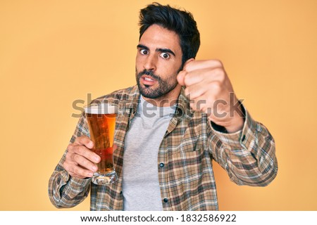 Handsome hispanic man with beard drinking a pint of beer annoyed and frustrated shouting with anger, yelling crazy with anger and hand raised 