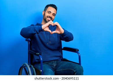 Handsome Hispanic Man With Beard Sitting On Wheelchair Smiling In Love Doing Heart Symbol Shape With Hands. Romantic Concept. 