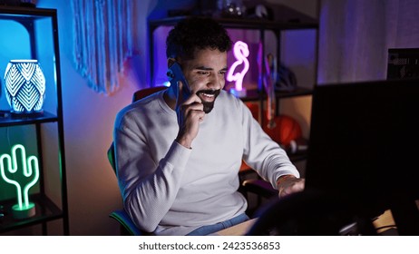 Handsome hispanic man with beard smiling in a colorful illuminated gaming room at night, engaged in a conversation on the phone - Powered by Shutterstock