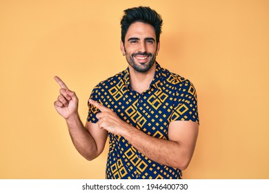 Handsome Hispanic Man With Beard Wearing 80s Disco Style Shirt Smiling And Looking At The Camera Pointing With Two Hands And Fingers To The Side. 