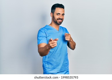 Handsome Hispanic Man With Beard Wearing Blue Male Nurse Uniform Pointing Fingers To Camera With Happy And Funny Face. Good Energy And Vibes. 