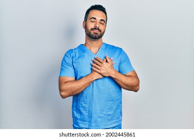 Handsome Hispanic Man With Beard Wearing Blue Male Nurse Uniform Smiling With Hands On Chest With Closed Eyes And Grateful Gesture On Face. Health Concept. 