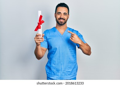 Handsome Hispanic Man With Beard Wearing Blue Male Nurse Uniform Holding Diploma Smiling Happy Pointing With Hand And Finger 