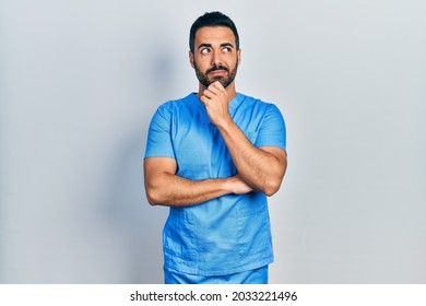 Handsome Hispanic Man With Beard Wearing Blue Male Nurse Uniform With Hand On Chin Thinking About Question, Pensive Expression. Smiling With Thoughtful Face. Doubt Concept. 