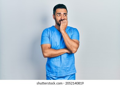 Handsome Hispanic Man With Beard Wearing Blue Male Nurse Uniform Looking Stressed And Nervous With Hands On Mouth Biting Nails. Anxiety Problem. 