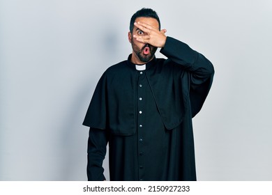 Handsome hispanic man with beard wearing catholic priest robe peeking in shock covering face and eyes with hand, looking through fingers with embarrassed expression. 