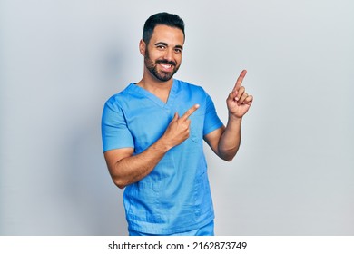 Handsome Hispanic Man With Beard Wearing Blue Male Nurse Uniform Smiling And Looking At The Camera Pointing With Two Hands And Fingers To The Side. 