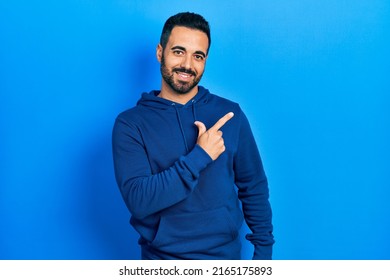 Un hombre hispano guapo con barba y sudadera casual sonriendo alegremente señalando con la mano y el dedo hacia el costado 