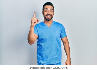 Handsome Hispanic Man With Beard Wearing Blue Male Nurse Uniform Pointing Finger Up With Successful Idea. Exited And Happy. Number One. 