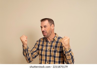 Handsome hispanic man with beard wearing checkered shirt over isolated beige background angry and crazy raising fists frustrated and furious while shouting with anger. Rage and aggressive concept - Shutterstock ID 2211875179