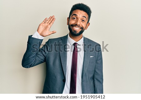 Handsome hispanic man with beard wearing business suit and tie waiving saying hello happy and smiling, friendly welcome gesture 