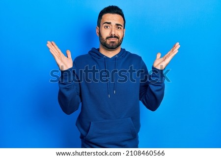Handsome hispanic man with beard wearing casual sweatshirt clueless and confused with open arms, no idea and doubtful face. 