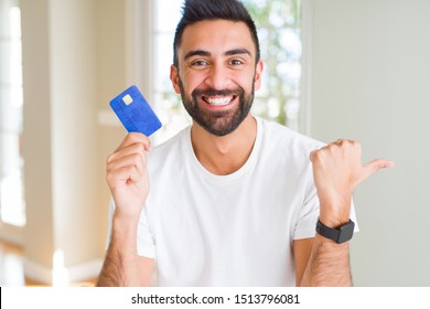 Handsome Hispanic Man Holding Credit Card Pointing And Showing With Thumb Up To The Side With Happy Face Smiling