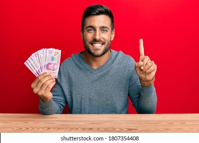 Handsome hispanic man holding mexican pesos smiling with an idea or question pointing finger with happy face, number one 