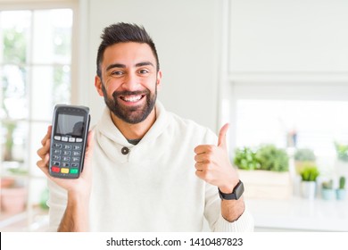 Handsome Hispanic Man Holding Point Of Sale Terminal Dataphone Happy With Big Smile Doing Ok Sign, Thumb Up With Fingers, Excellent Sign