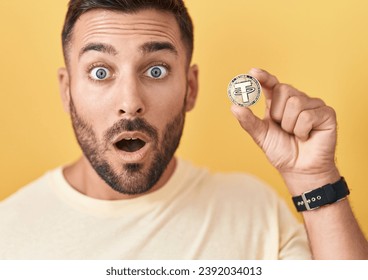 Handsome hispanic man holding tether cryptocurrency coin scared and amazed with open mouth for surprise, disbelief face 