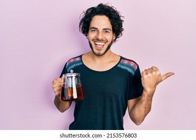 Handsome Hispanic Man Holding Traditional Tea Pot Pointing Thumb Up To The Side Smiling Happy With Open Mouth 