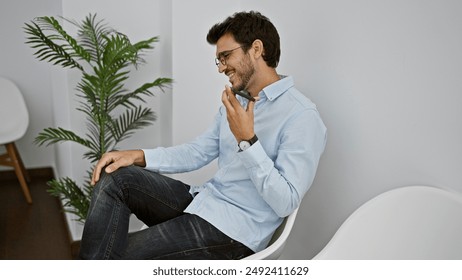 Handsome hispanic man laughing while listening to a voice message in a modern waiting room.