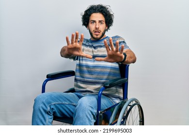 Handsome Hispanic Man Sitting On Wheelchair Afraid And Terrified With Fear Expression Stop Gesture With Hands, Shouting In Shock. Panic Concept. 