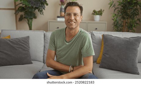 Handsome hispanic man sitting on a sofa in a cozy living room, smiling confidently towards the camera, surrounded by soft cushions and green plants, interior of a modern home - Powered by Shutterstock