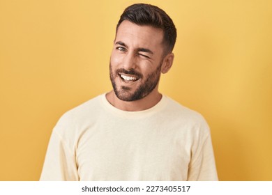 Handsome hispanic man standing over yellow background winking looking at the camera with sexy expression, cheerful and happy face. 