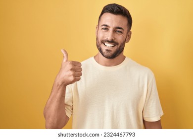 Handsome hispanic man standing over yellow background doing happy thumbs up gesture with hand. approving expression looking at the camera showing success.  - Powered by Shutterstock