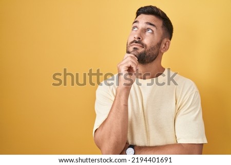 Handsome hispanic man standing over yellow background with hand on chin thinking about question, pensive expression. smiling with thoughtful face. doubt concept. 
