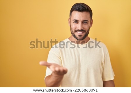 Handsome hispanic man standing over yellow background smiling cheerful offering palm hand giving assistance and acceptance. 