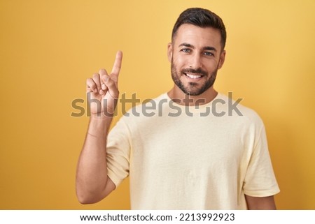 Handsome hispanic man standing over yellow background showing and pointing up with finger number one while smiling confident and happy. 