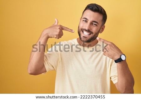 Handsome hispanic man standing over yellow background smiling cheerful showing and pointing with fingers teeth and mouth. dental health concept. 