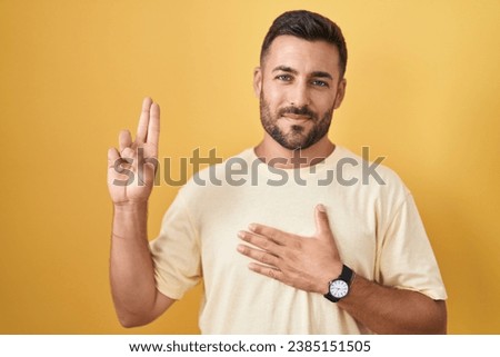 Handsome hispanic man standing over yellow background smiling swearing with hand on chest and fingers up, making a loyalty promise oath 