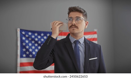 Handsome hispanic man in suit using smartphone in office with american flag