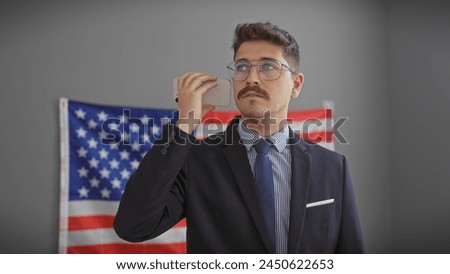 Handsome hispanic man in suit using smartphone in office with american flag