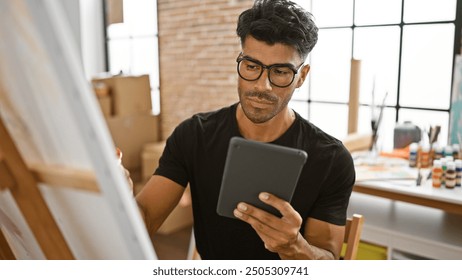 Handsome hispanic man using tablet in art studio with easel and paints - Powered by Shutterstock