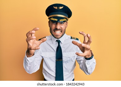 Handsome Hispanic Man Wearing Airplane Pilot Uniform Shouting Frustrated With Rage, Hands Trying To Strangle, Yelling Mad 