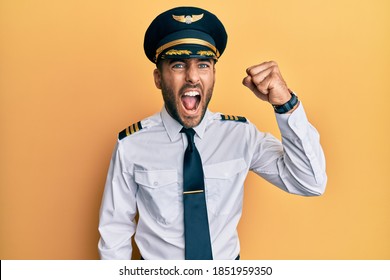 Handsome Hispanic Man Wearing Airplane Pilot Uniform Angry And Mad Raising Fist Frustrated And Furious While Shouting With Anger. Rage And Aggressive Concept. 