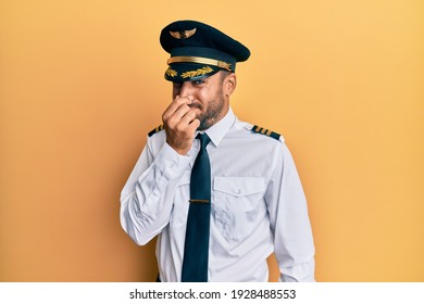 Handsome Hispanic Man Wearing Airplane Pilot Uniform Smelling Something Stinky And Disgusting, Intolerable Smell, Holding Breath With Fingers On Nose. Bad Smell 